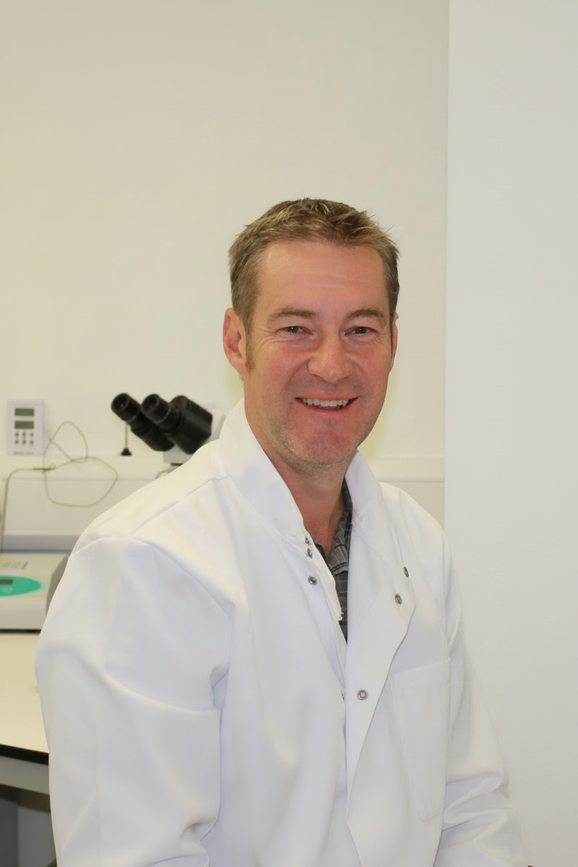 A smiling man in a white lab coat standing indoors next to laboratory equipment.